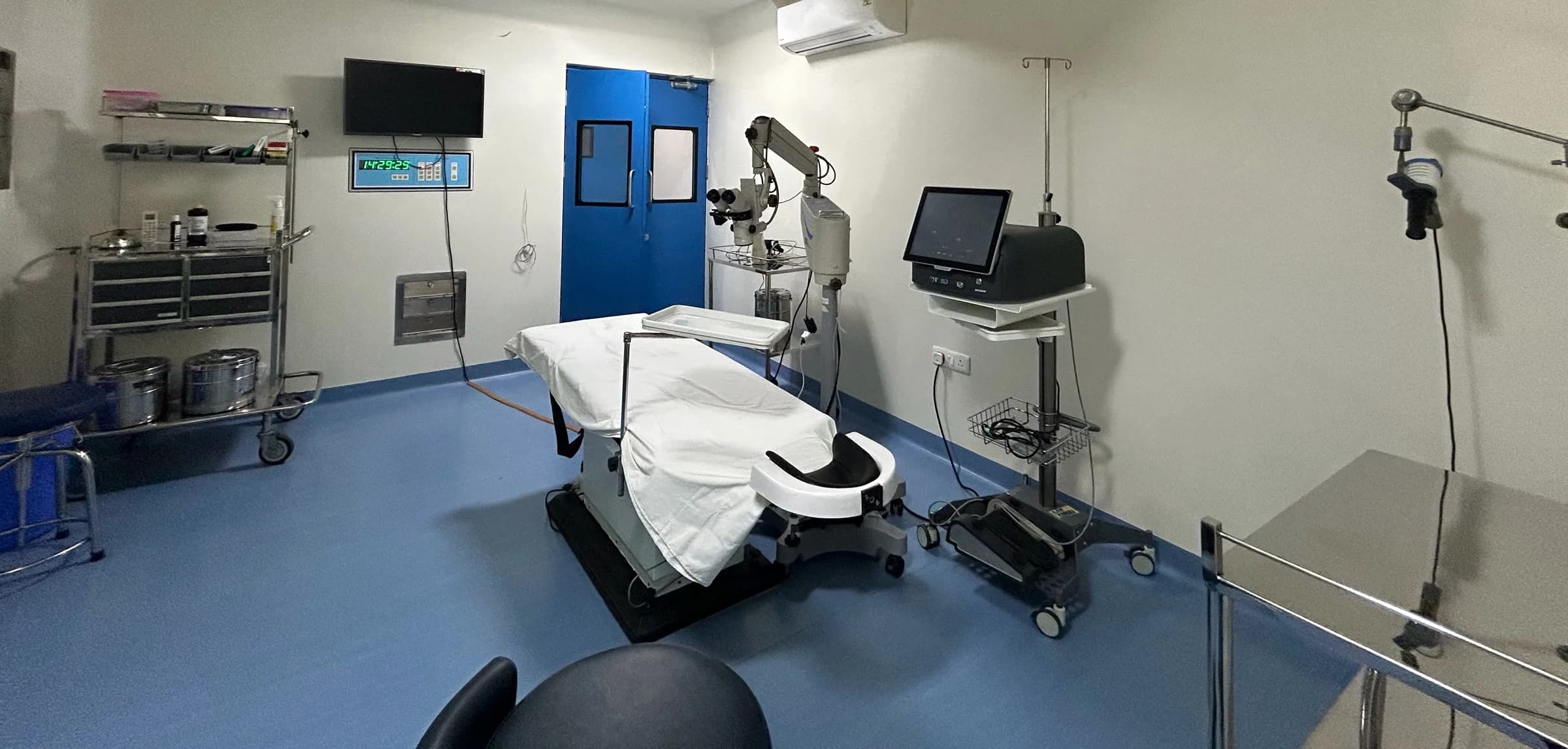 An operation theatre with an operating table, operating microscope, surgical console and a clock panel on the wall