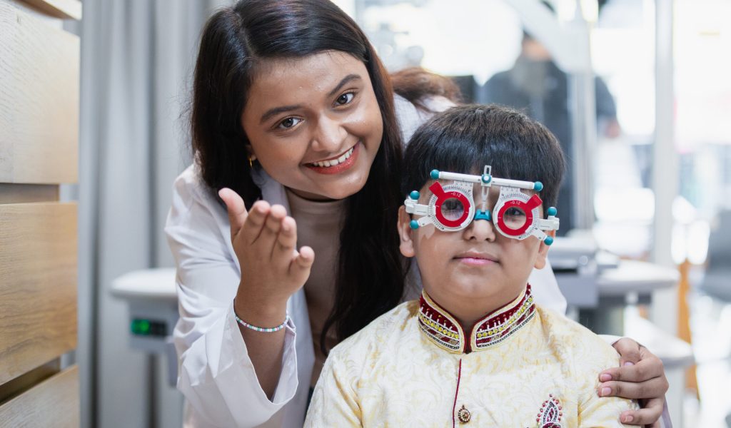 Happy Indian child boy with women eye specialist examining eyesight modern ophthalmology equipment in clinic. Patient kid male checkup iris examines ophthalmological hospital. measure eyeglasses.