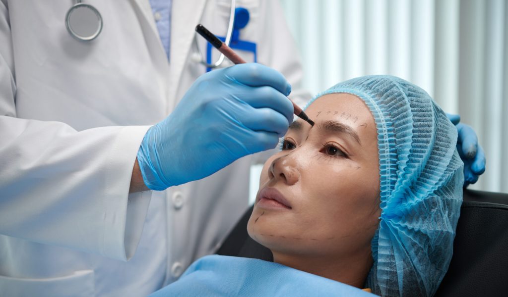 Plastic surgeon marking areas on face of young Asian woman before doing procedure
